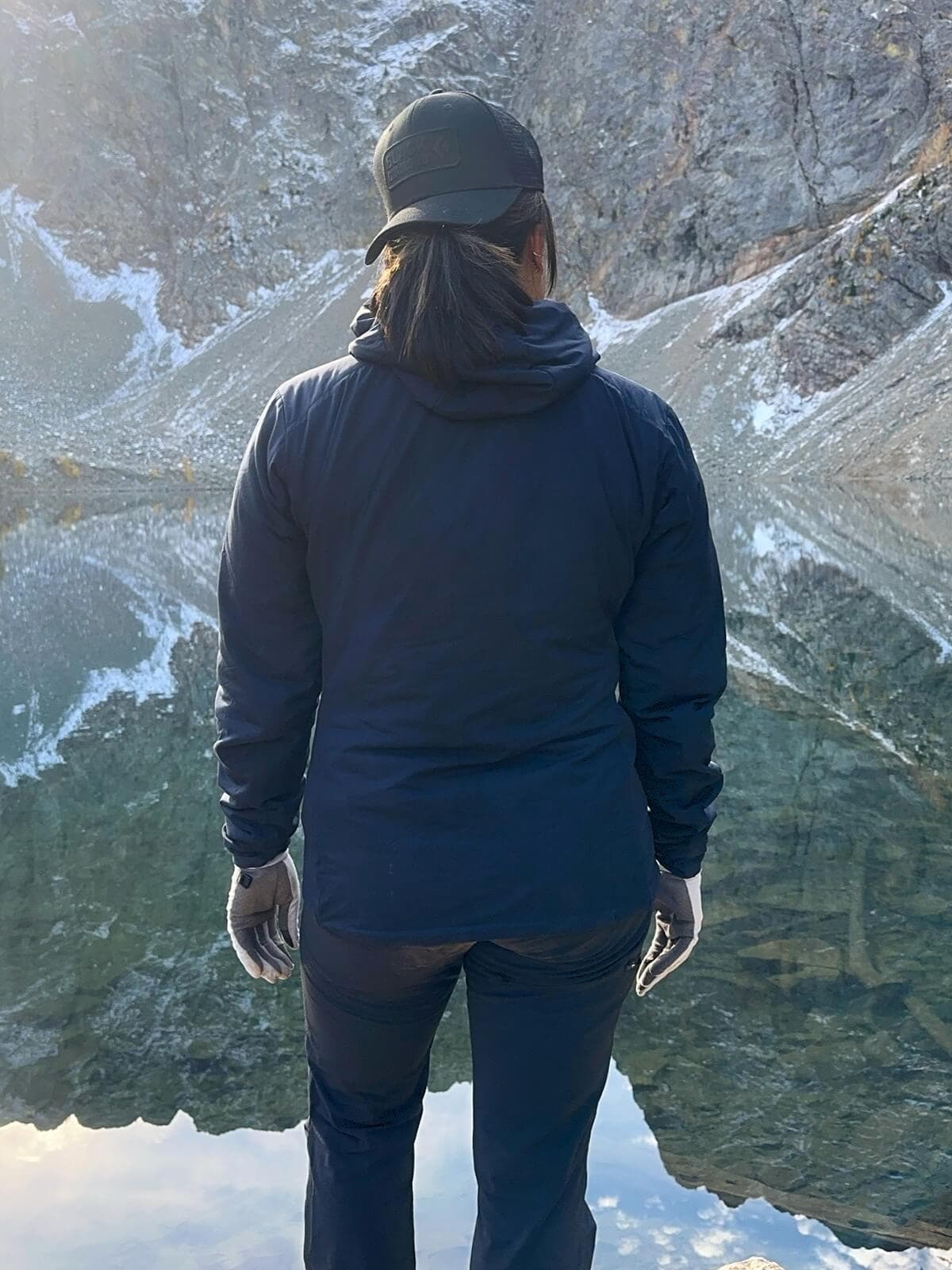 A hiker wearing a fitted hiking jacket, black pants, and gray gloves stands with her back to the camera beside a clear alpine lake. Steep rocky mountains with patches of snow reflect in the still water behind her.